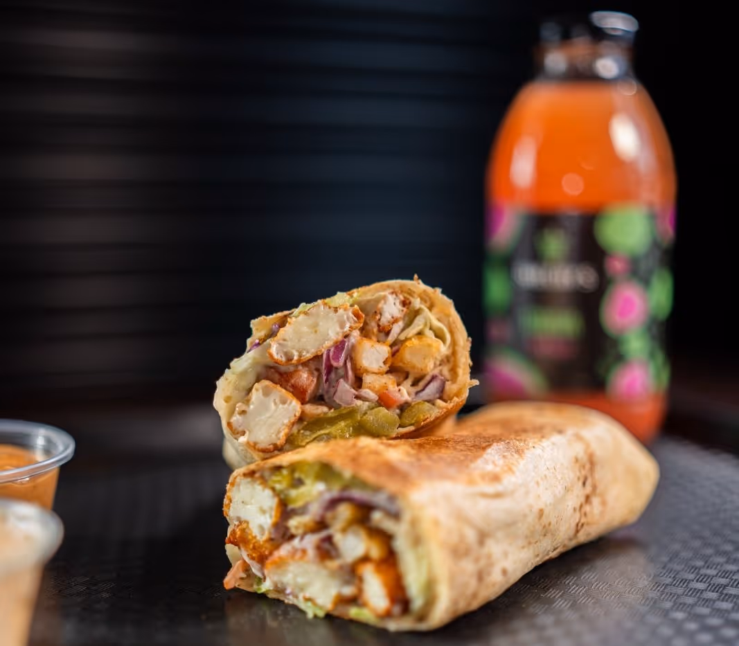 Close-up of a cut wrap sandwich filled with fried paneer, vegetables, and sauces on a black surface with a bottle of orange drink in the background.