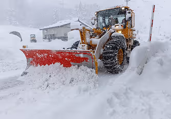Yellow snow plow clearing heavy snow on a street during a snowstorm near houses.