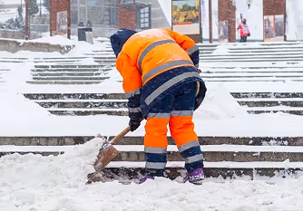 Worker in orange reflective gear shoveling snow on a stairway outdoors in winter.
