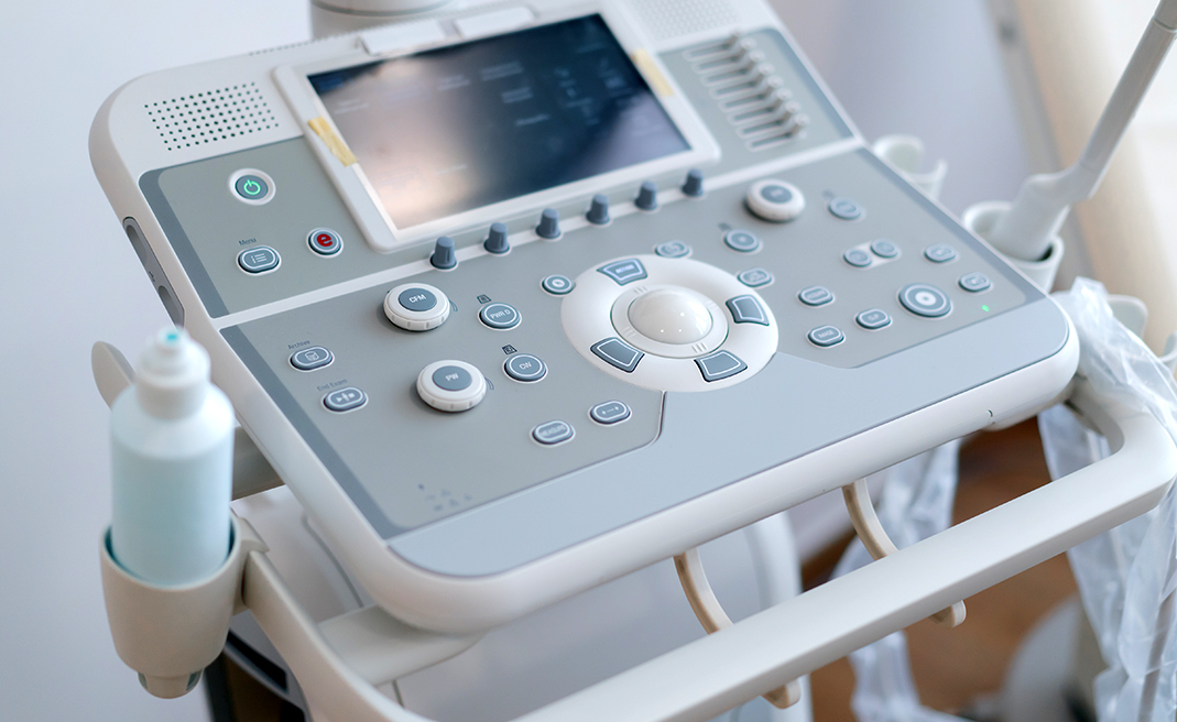 Close-up of a gray ultrasound machine console with buttons, screen, and gel bottle.