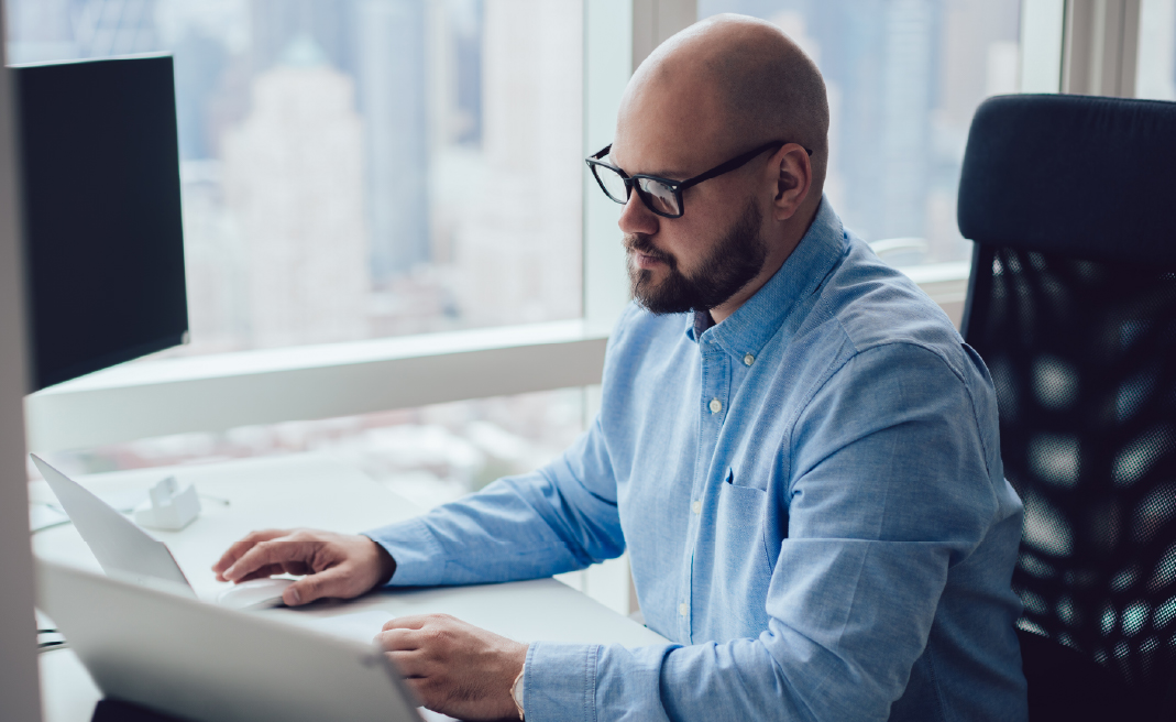 Bearded man in glasses and blue shirt working on laptop in office with city view window.