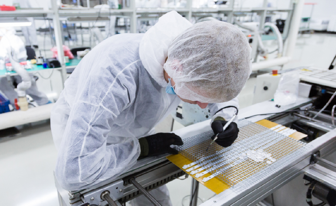 Person in protective gear inspecting or assembling medical device parts on a clean factory line.