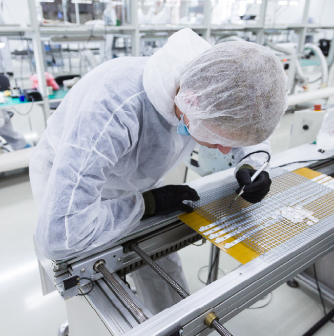 Person in protective gear inspecting small electronic components on a workbench in a lab.