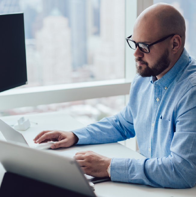 Man with glasses and blue shirt working on a laptop at a desk near a large window.