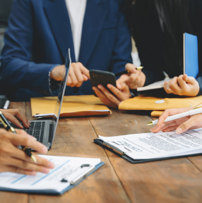 People in business attire reviewing documents, using a laptop and smartphone at a wooden table.