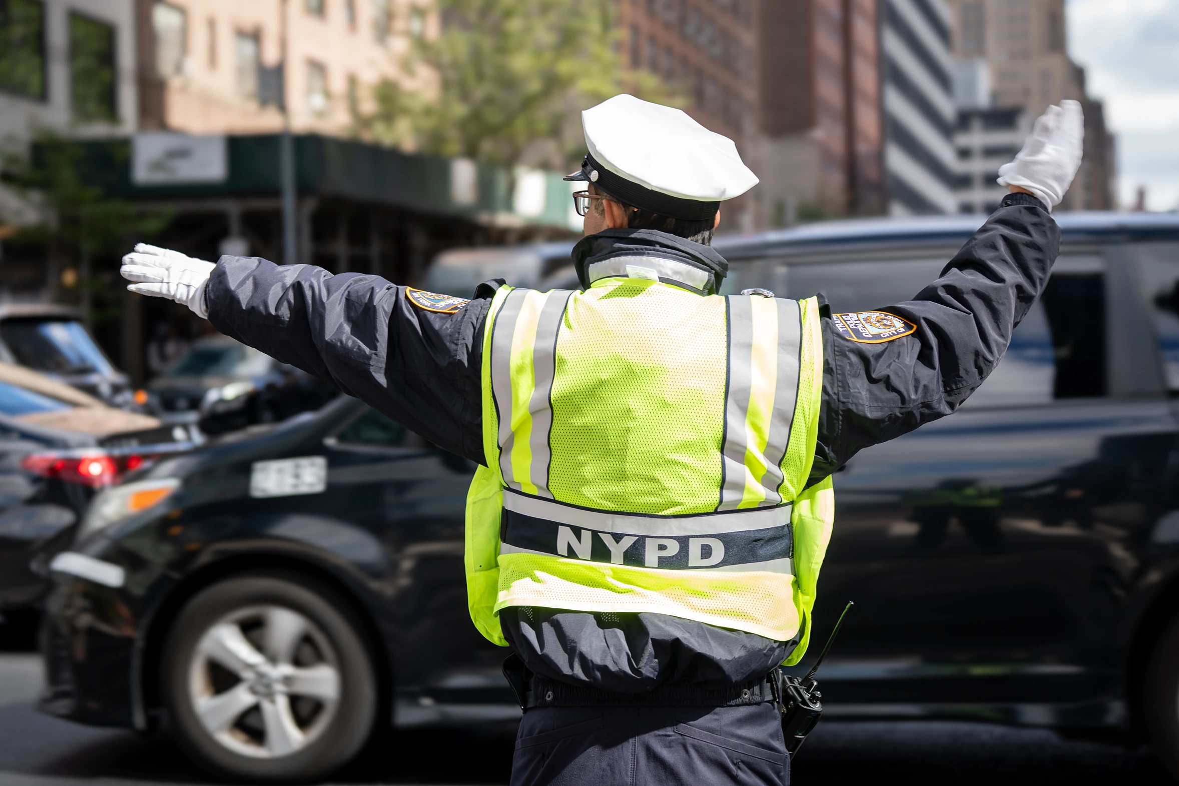 NYPD officer wearing a white cap and reflective vest directing traffic with arms outstretched on a city street.