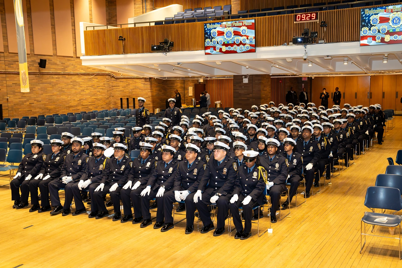 Large group of police officers in formal uniforms and white gloves seated in rows inside an auditorium with wooden floors and walls.