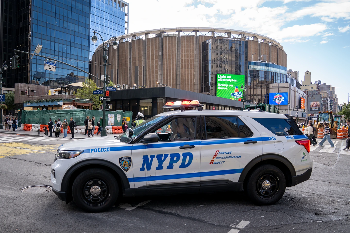 NYPD police SUV with lights on driving on a city street near a large round building and pedestrians crossing at the intersection.