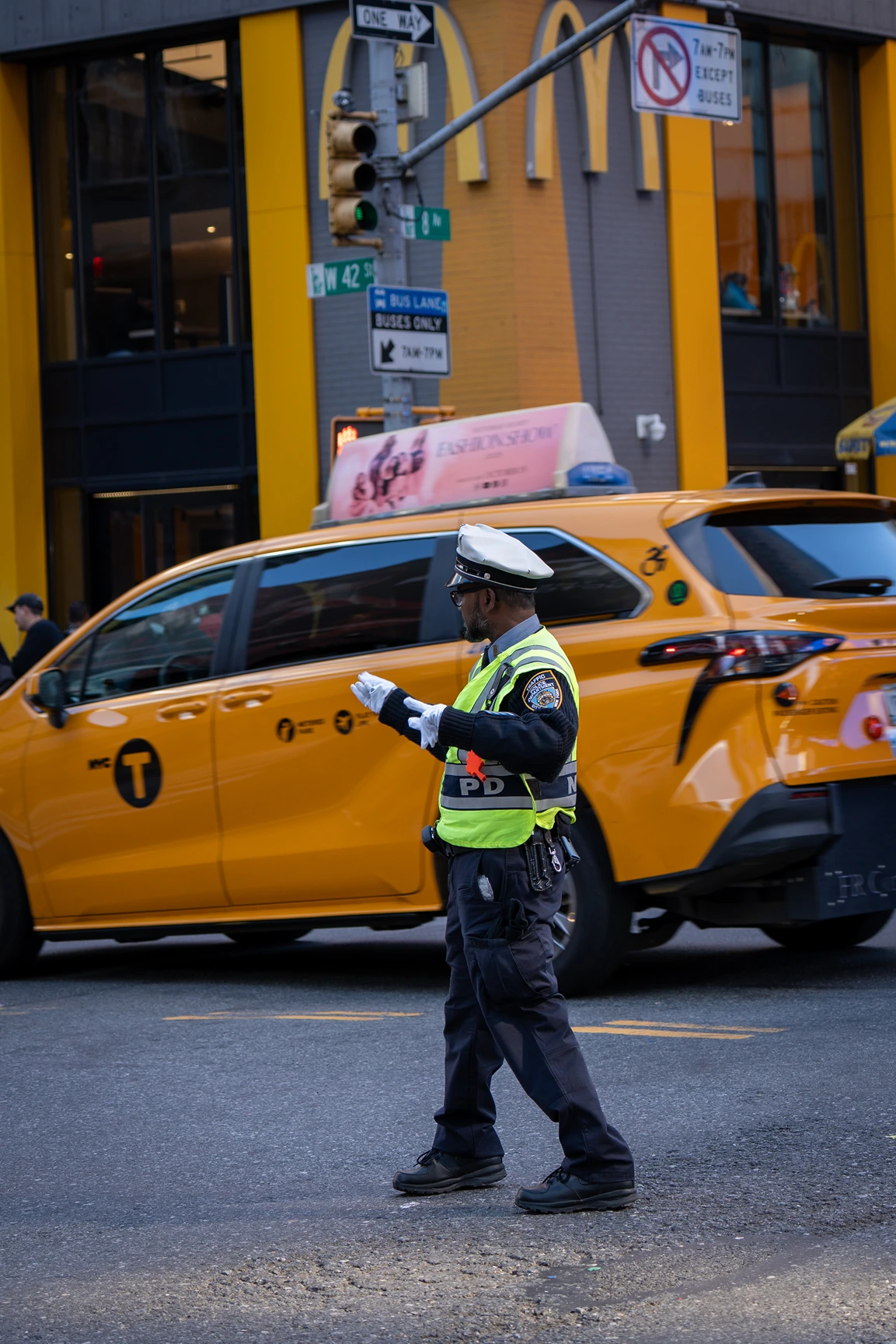 NYPD traffic officer directing vehicles in front of a yellow NYC taxi at an intersection with street signs.