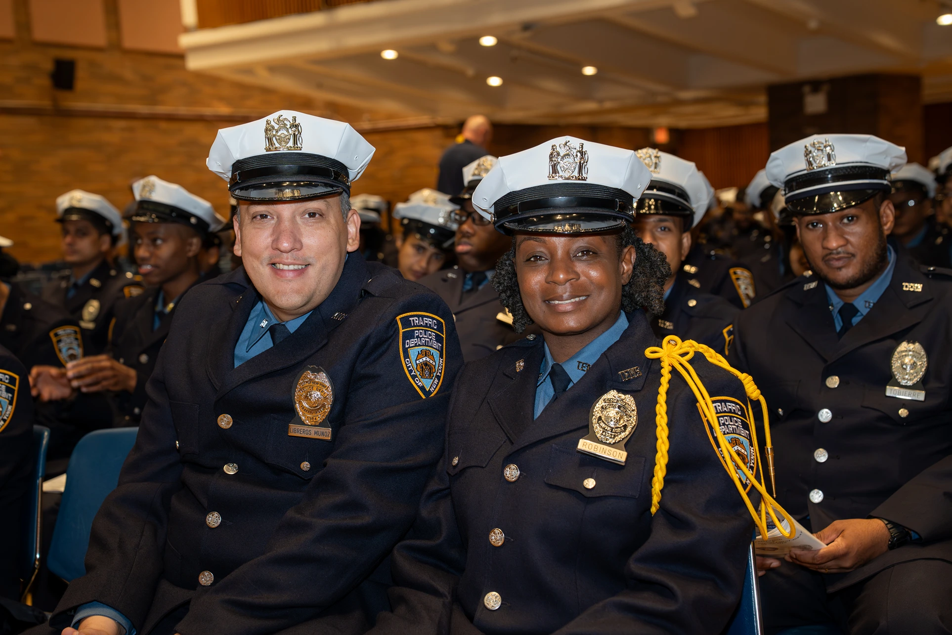 Smiling uniformed traffic police officers seated indoors during an event, wearing white hats with badges and navy blue coats.