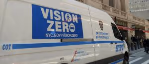 White NYPD van with large blue Vision Zero logo and text promoting NYC.gov/VisionZero on a city street.