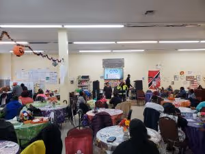 Community meeting in a large room with people seated at tables listening to speakers near a presentation screen displaying the number 1070.