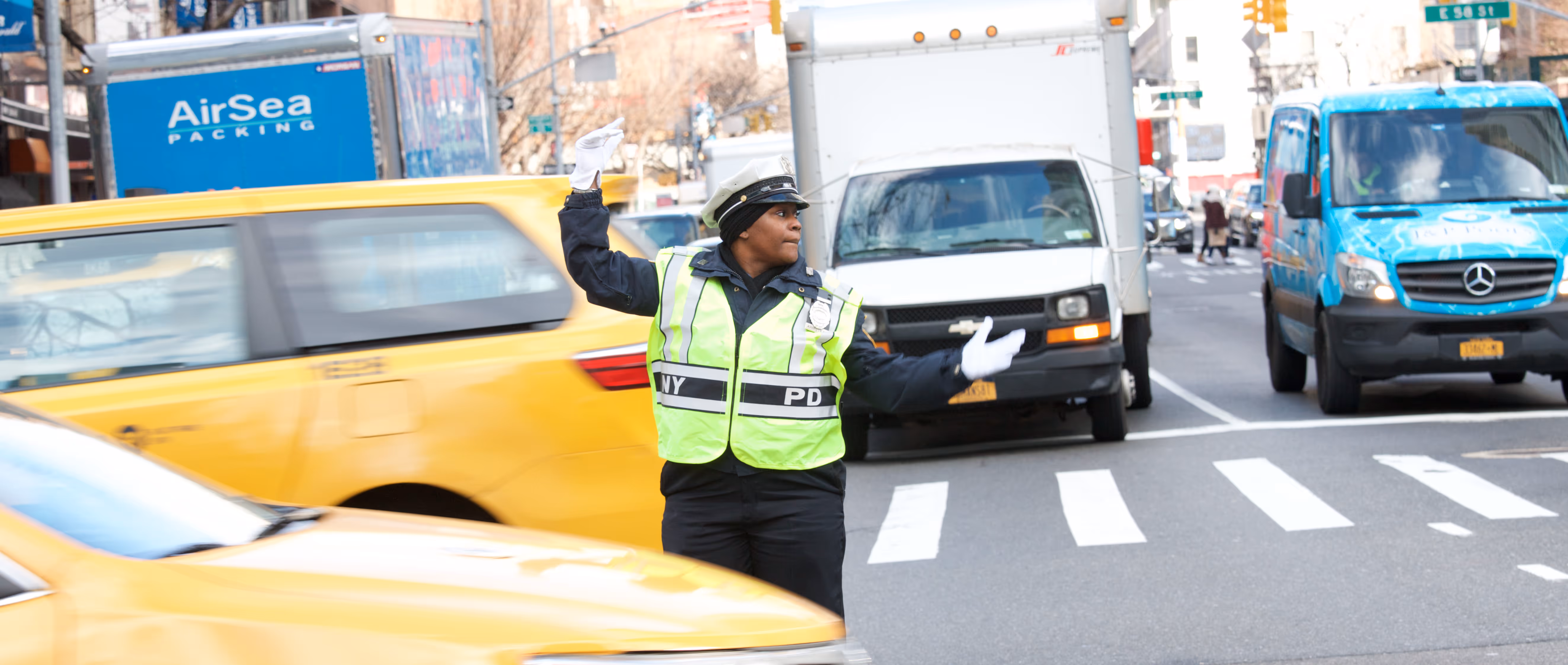 A NYPD officer wearing a reflective vest directs traffic on a busy city street with yellow taxis and delivery vans.