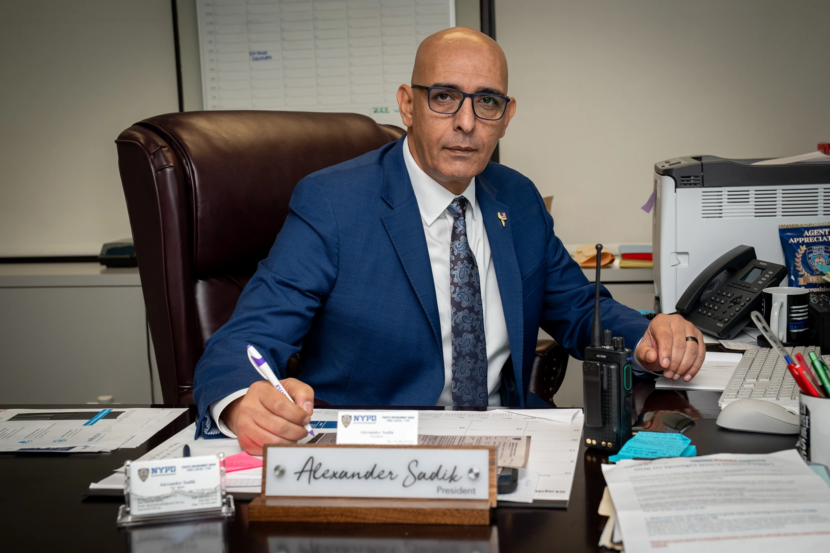 Man in a blue suit and glasses sitting at a desk with a nameplate reading Alexander Sadik, President, writing with a pen.