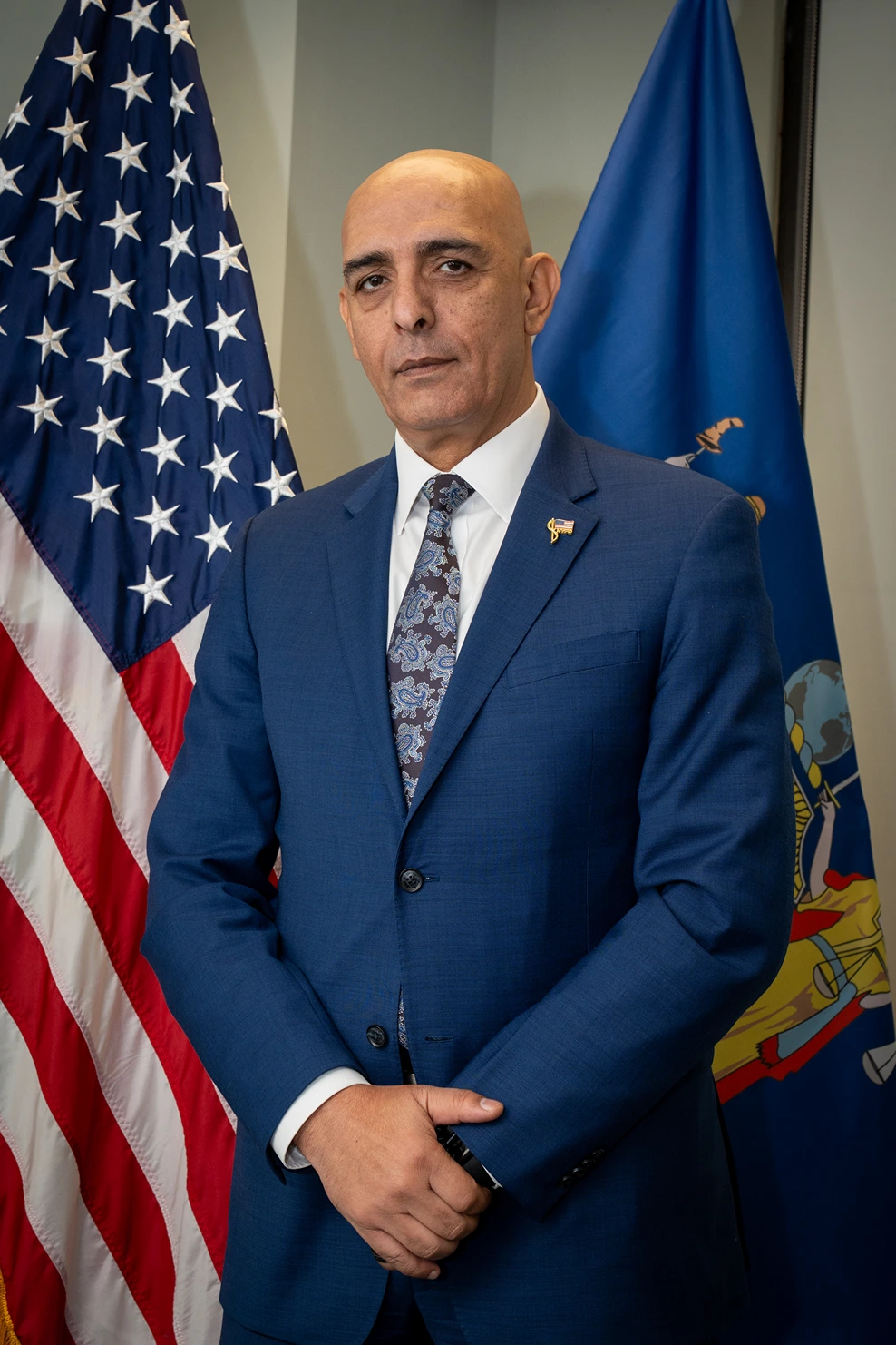 Man in a blue suit with a patterned tie standing with hands clasped in front, American and New York state flags in the background.