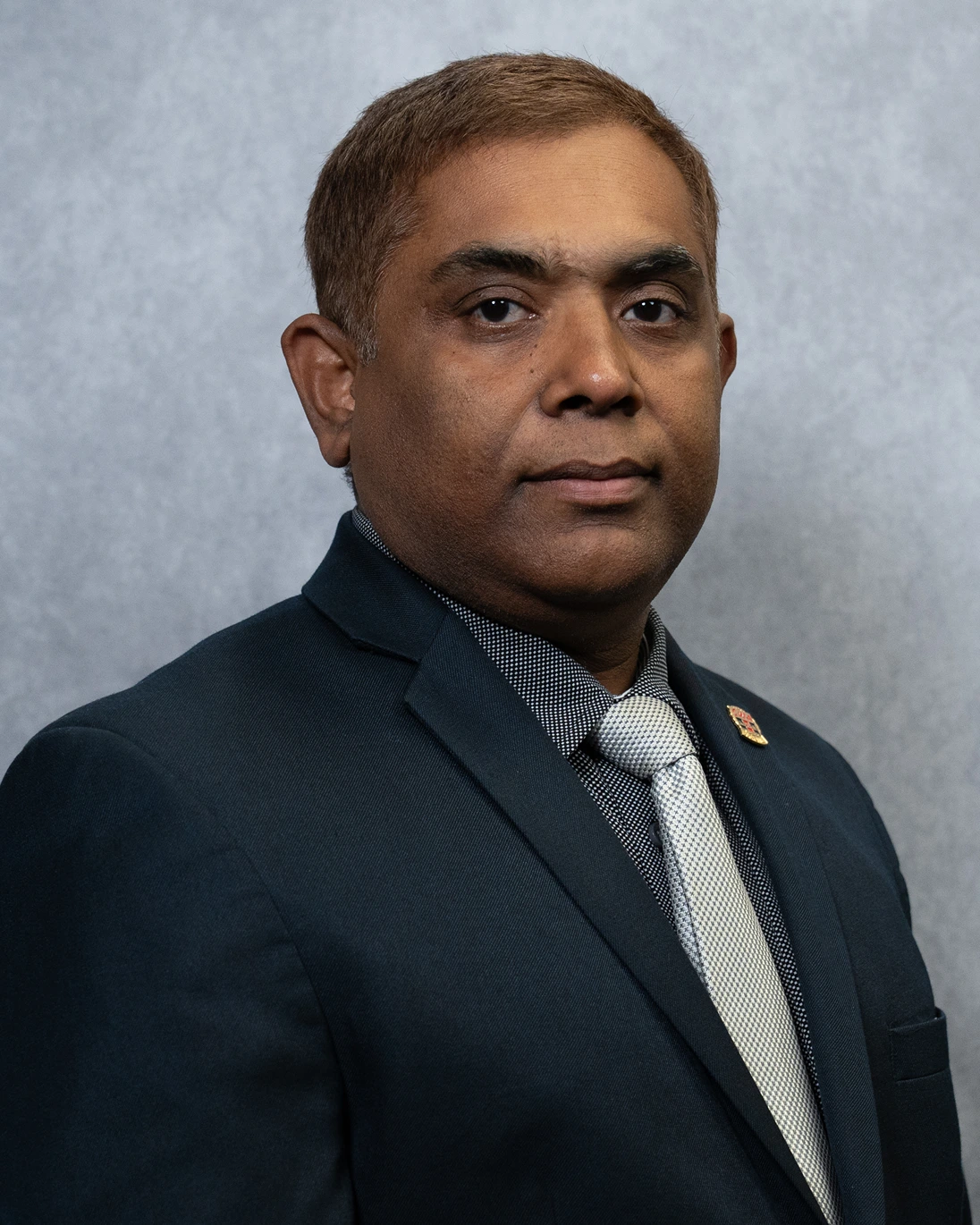 Portrait of a man in a dark suit, patterned shirt, and light-colored tie against a gray background.
