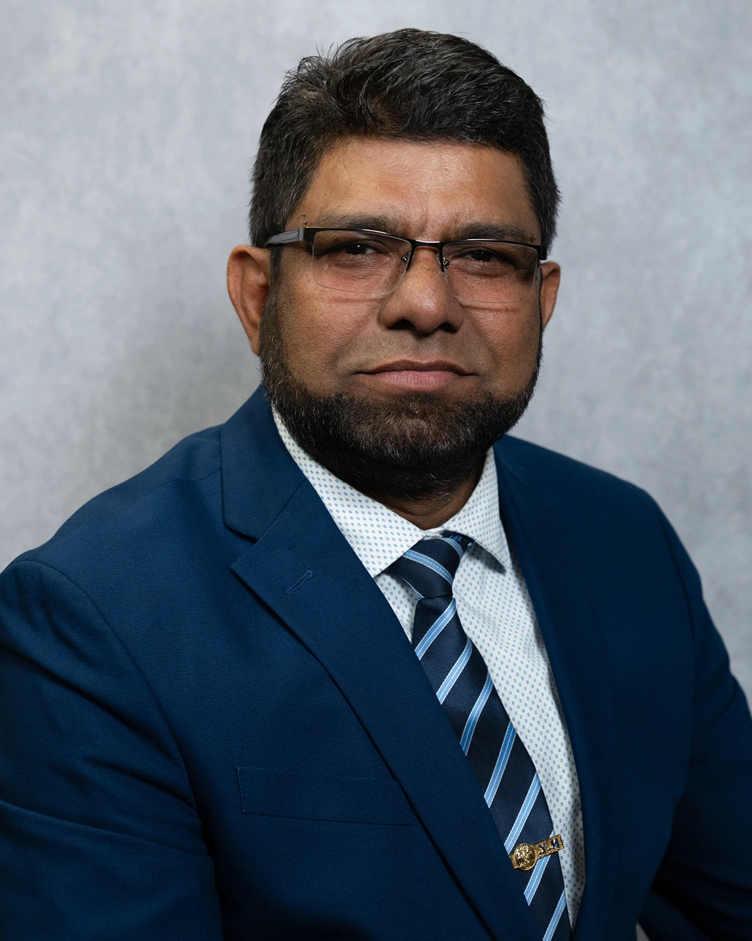 Man wearing glasses, a blue suit, white patterned shirt, and striped blue tie, posing against a gray background.