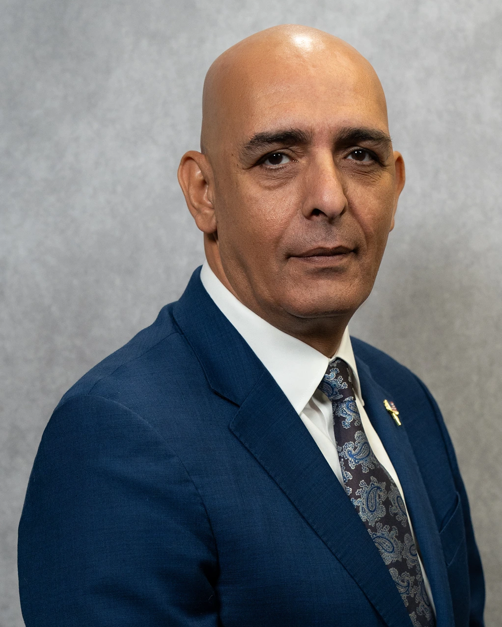 Portrait of a bald man wearing a blue suit, white shirt, and patterned tie, posed against a gray background.