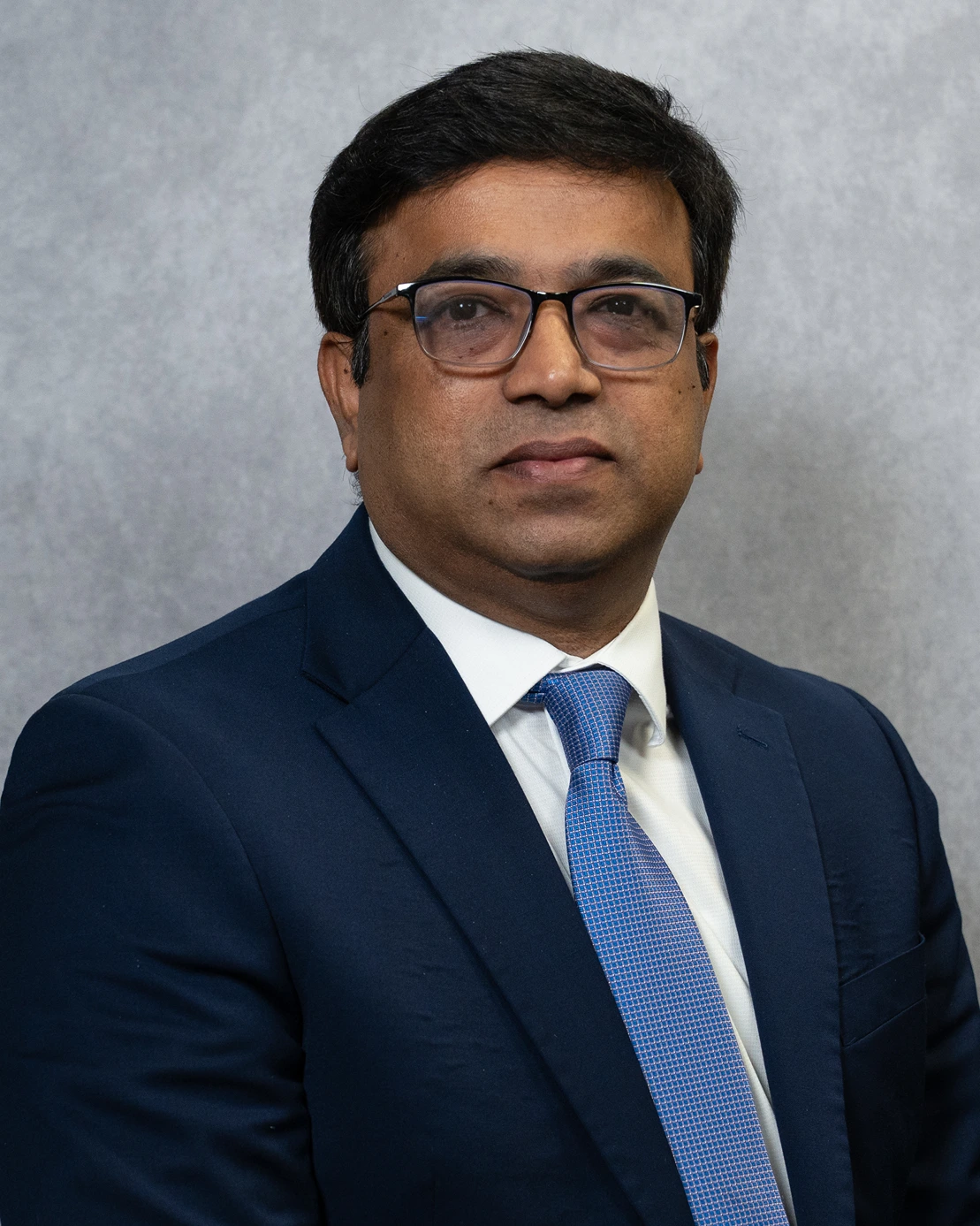 Man with glasses wearing a dark blue suit, white shirt, and light blue tie in front of a gray background.