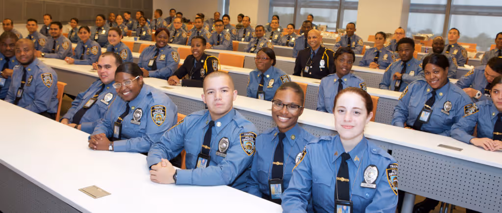 Group of uniformed police officers in a classroom, seated and facing forward, smiling.