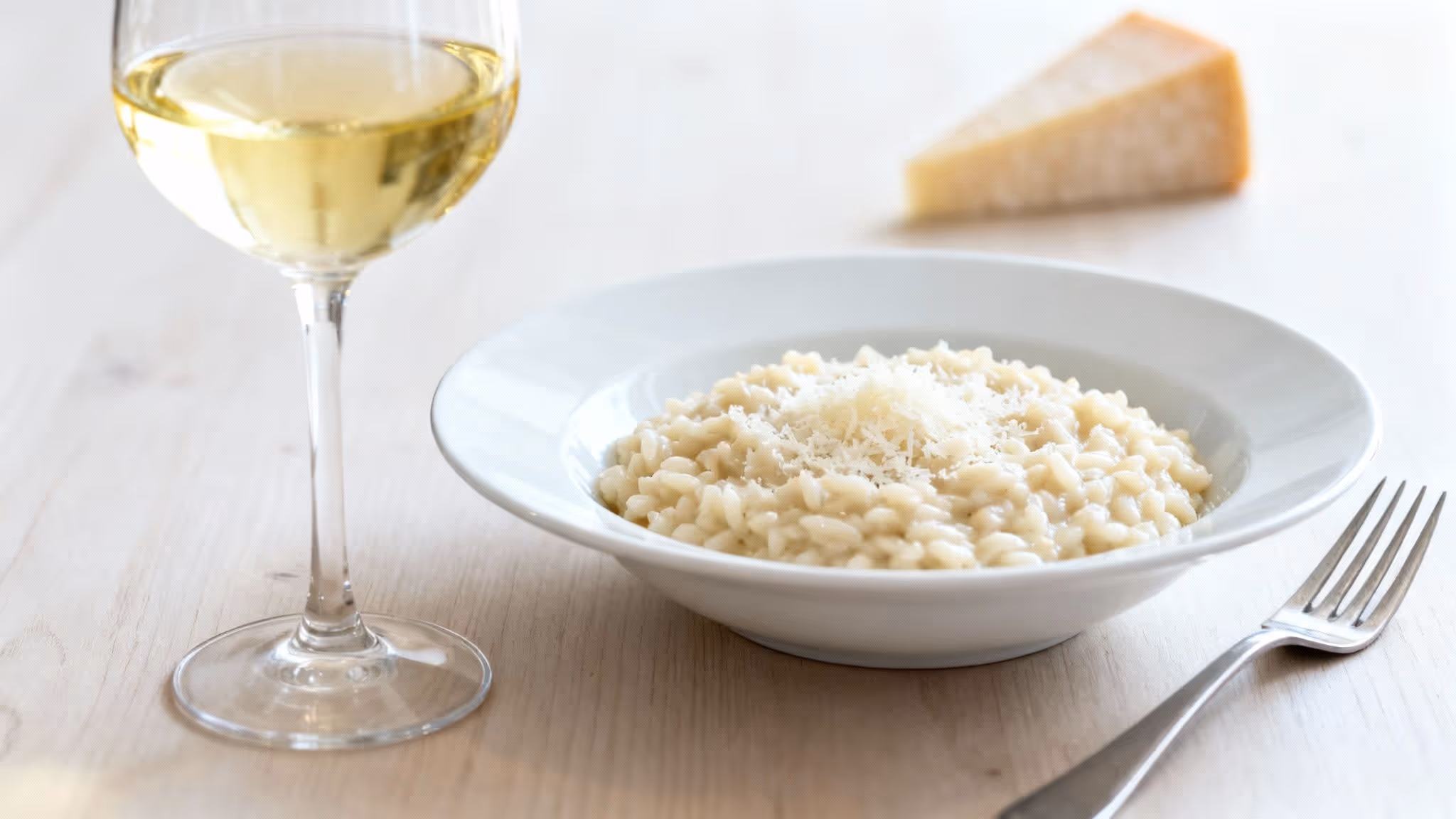 A creamy plate of risotto with grated Parmesan cheese, a glass of white wine, and a fork.