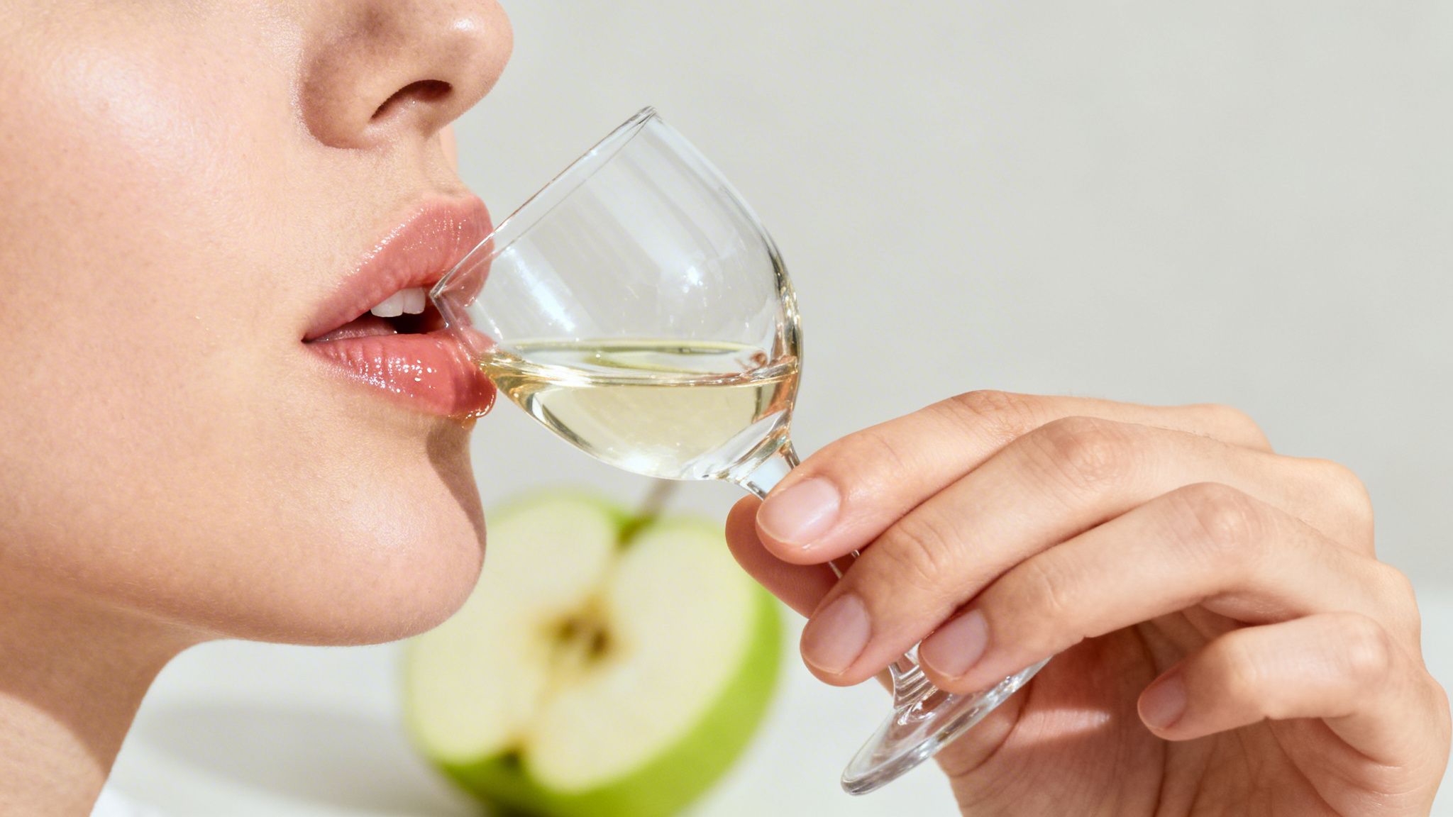 Close-up of a person with glossy lips tasting a light-colored beverage from a small glass, with a green apple in the background.