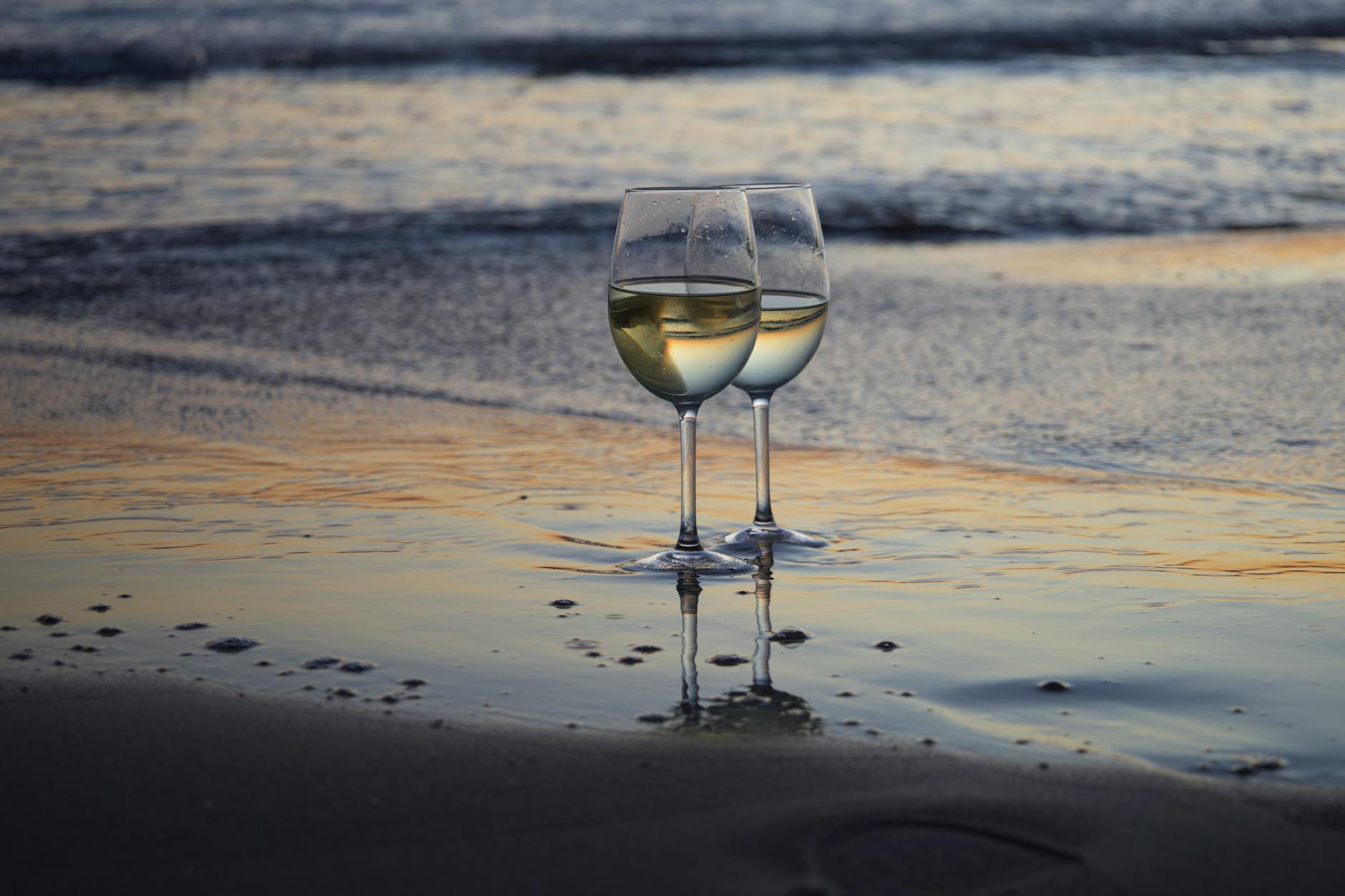 Two glasses of white wine on a beach during a serene sunset, reflecting warm colors.