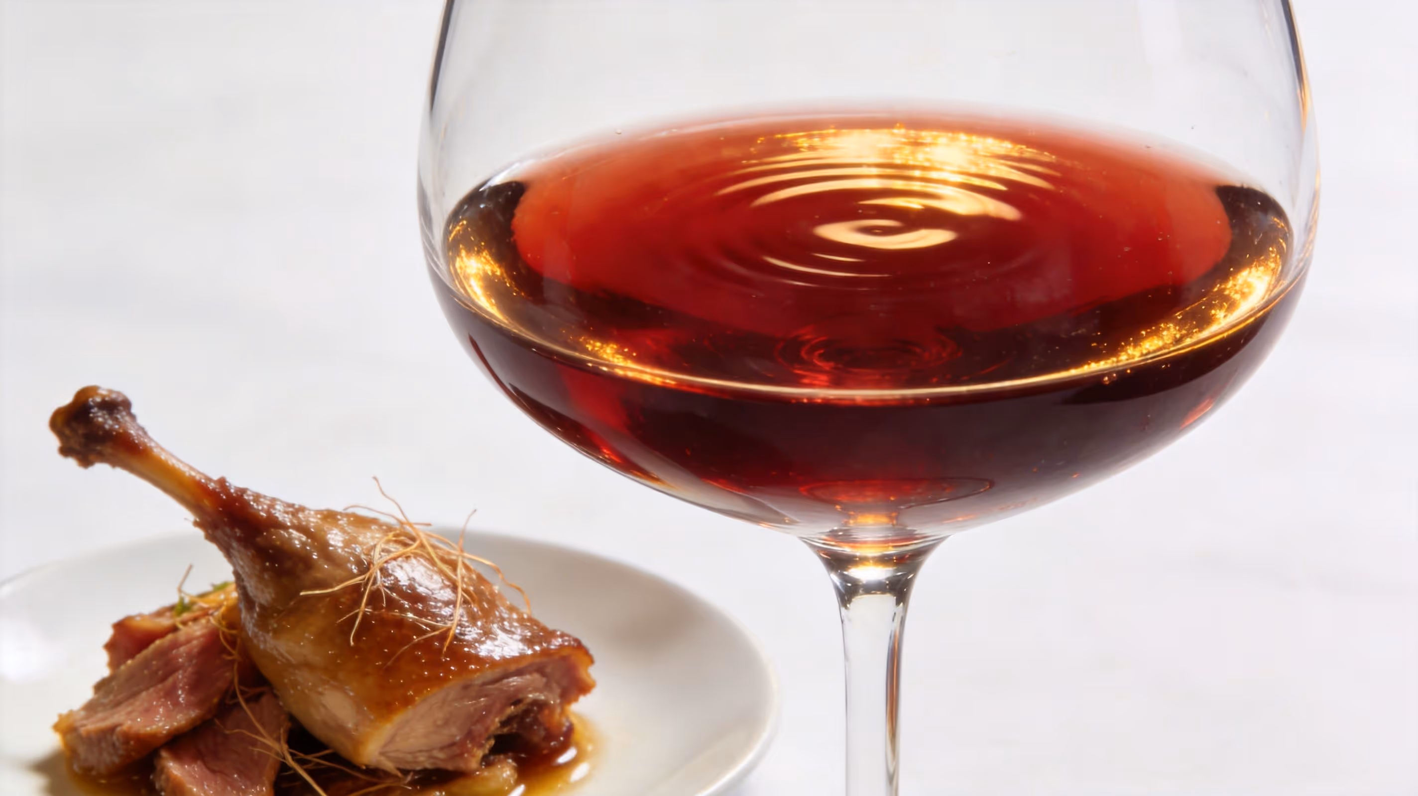 A close-up of a glass of red wine with ripples next to a plate of roasted duck.