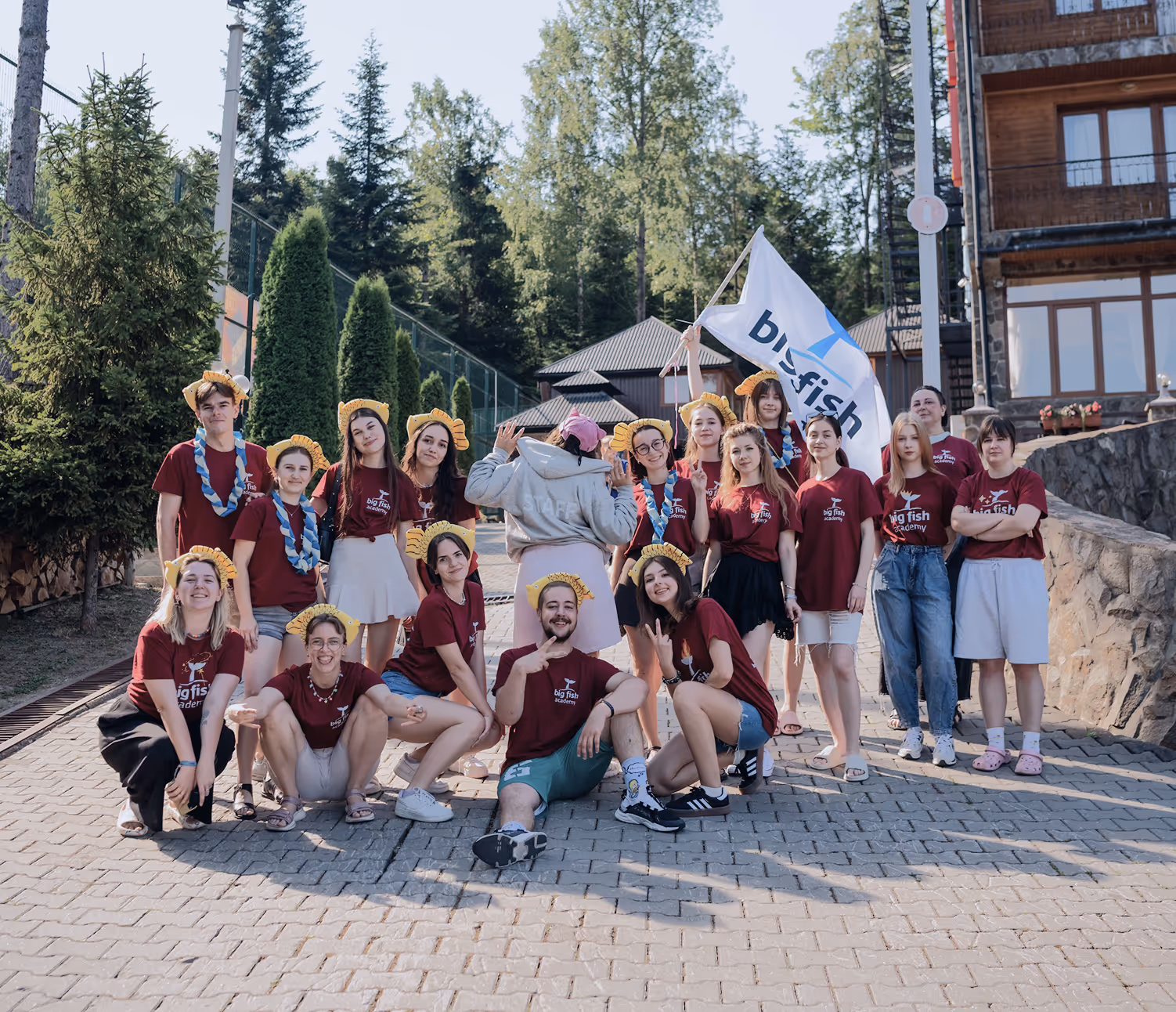 Група молодих людей у червоних футболках big fish academy позує на вулиці на тлі дерев і будівель, одна людина тримає прапор з написом big fish.