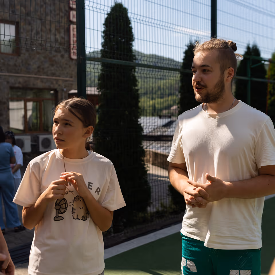 Молоді люди у повсякденному одязі розмовляють на тлі сітчастого паркану з деревами і будівлею позаду.