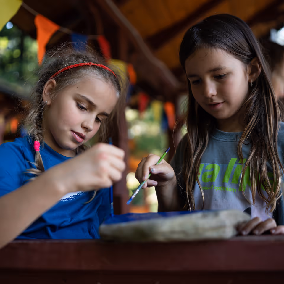 Дві дівчинки малюють кистями на камені за столом у приміщенні з яскравими прапорцями на задньому плані.