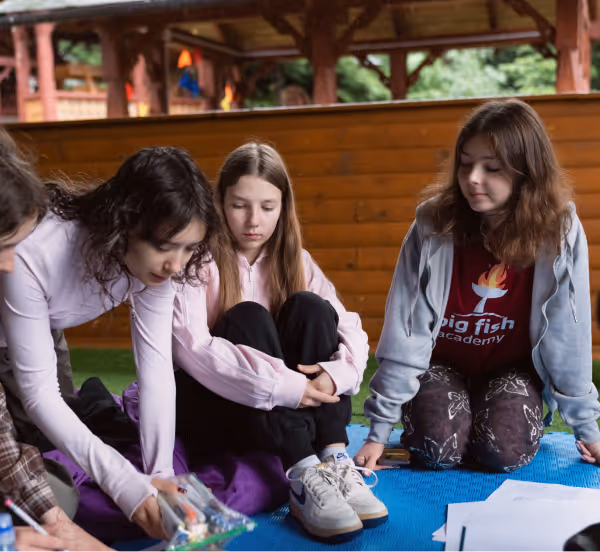Три дівчини сидять на килимку в критому просторі, одна з них складає руки на колінах, інша дивиться на предмет у руках третьої.