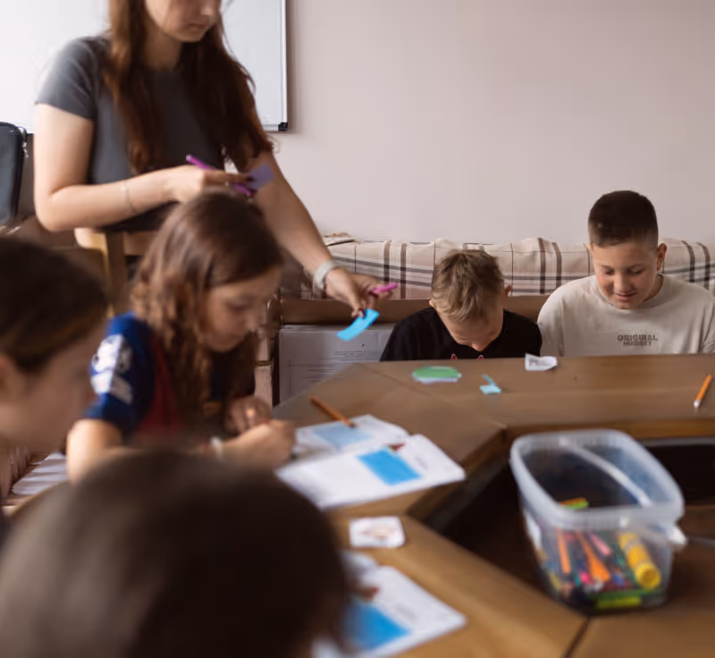 Вчителька допомагає дітям за круглим столом із розмальовками та олівцями.