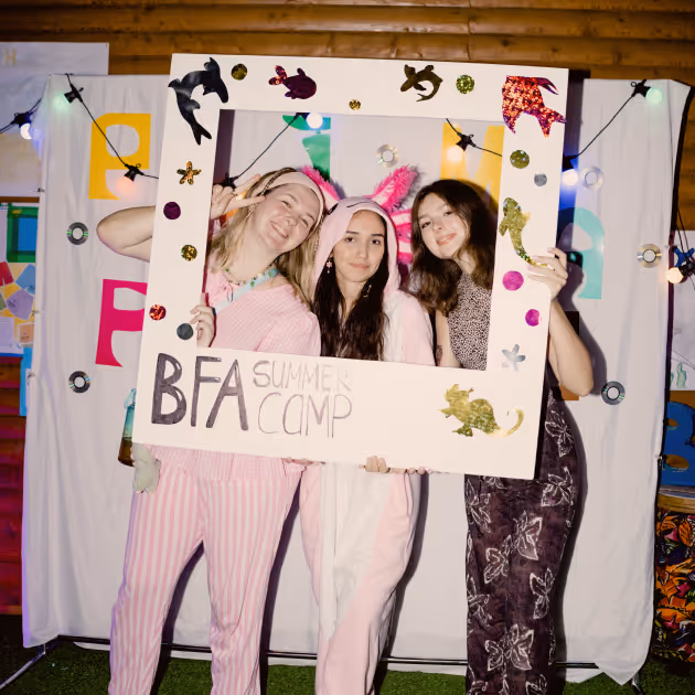 Три дівчини в піжамах позують, тримаючи рамку з написом BFA Summer Camp на вечірці з декоративним освітленням.