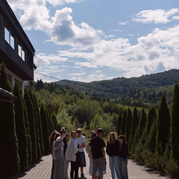 Група людей стоїть на мощеній доріжці між рядами високих зелених кущів на фоні лісистих пагорбів під блакитним небом з хмарами.