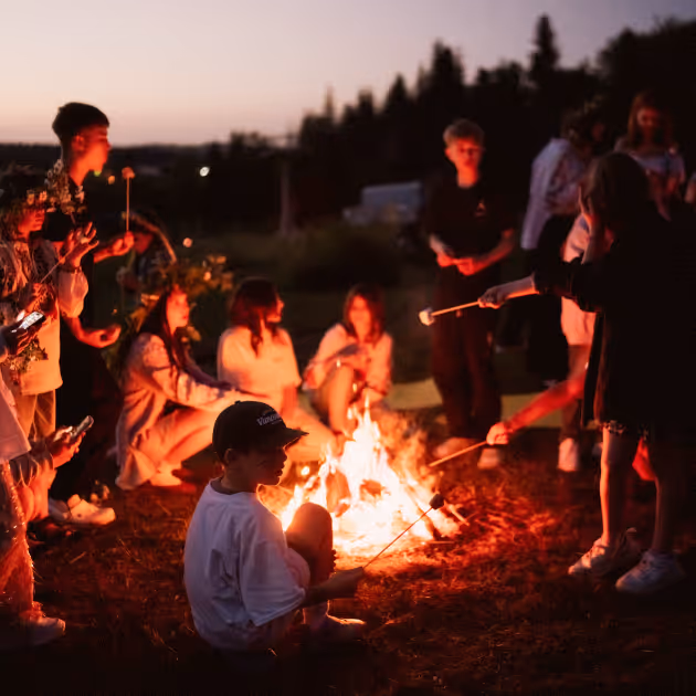 Група людей, в основному дітей та підлітків, сидять і стоять навколо багаття на заході сонця і підсмажують маршмеллоу на паличках.