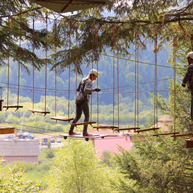 Жінка в захисному шоломі та спорядженні йде по мотузковому місту серед дерев у лісі на фоні зелених пагорбів.