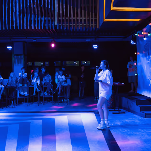 Молода жінка співає в мікрофон на сцені з синім освітленням, а аудиторія уважно слухає на фоні.