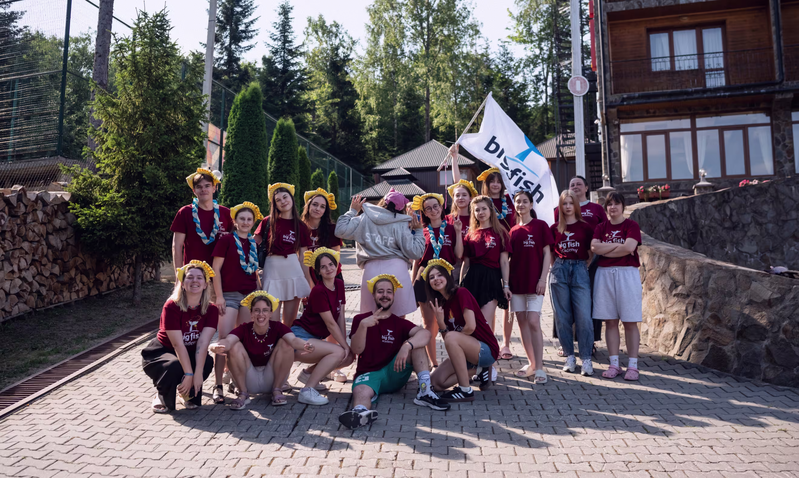 Група молодих людей у бордових футболках з написом Big Fish Academy позує на вулиці біля будинку, деякі з них у жовтих головних уборах, а одна людина тримає прапор Big Fish.