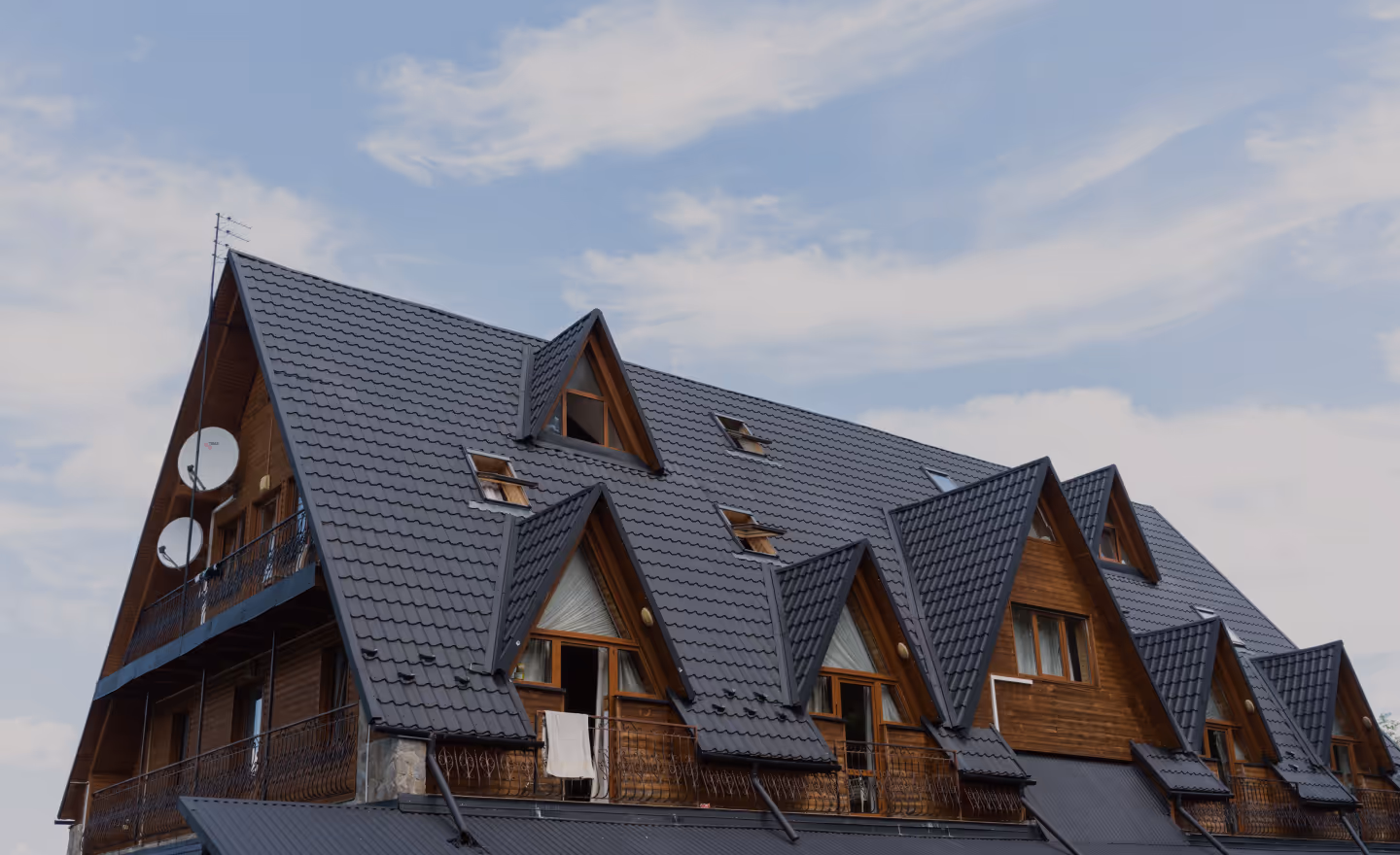 Великий дерев'яний будинок з металевою даховою черепицею та кількома мансардними вікнами під хмарним небом.