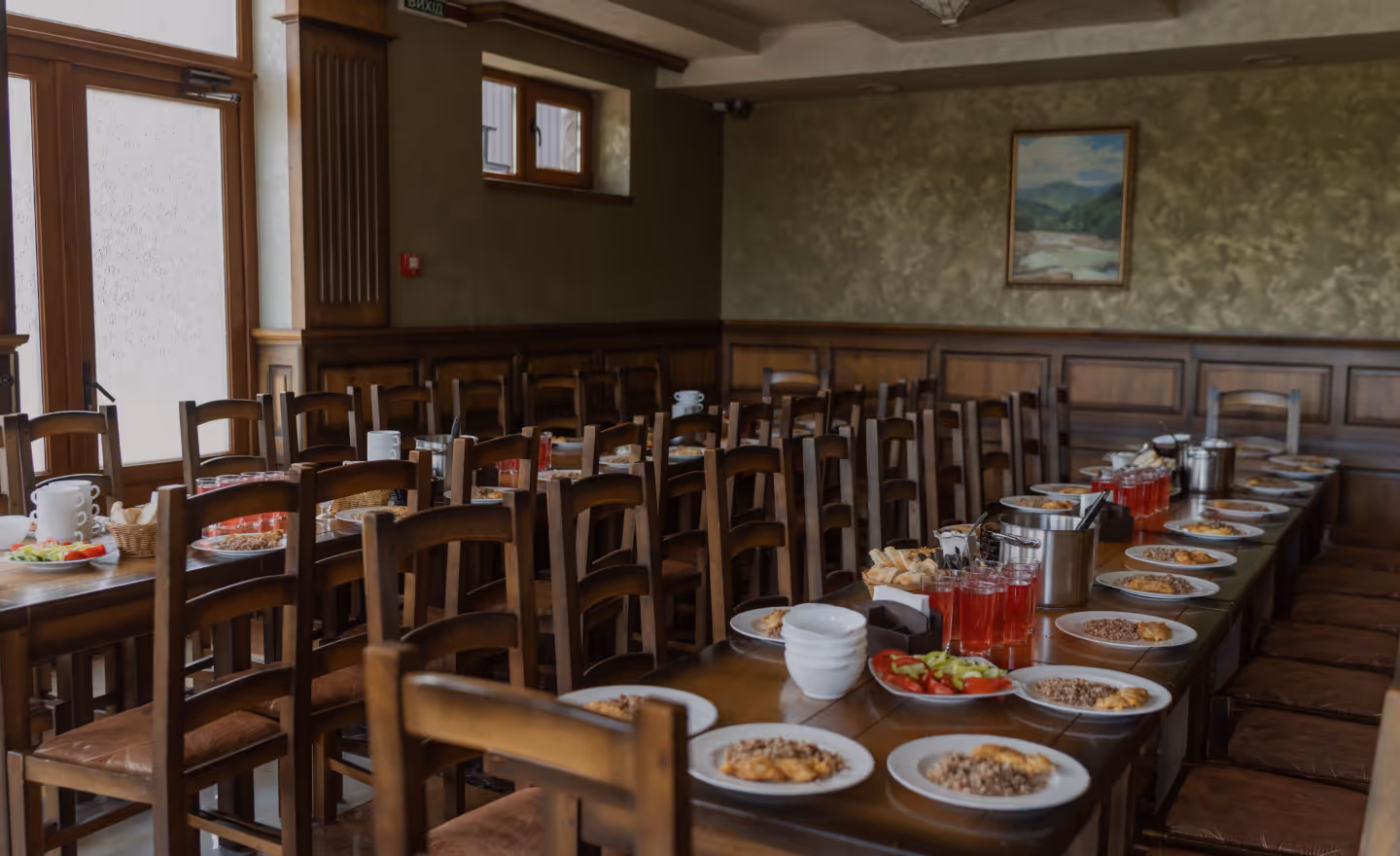 Зала ресторану з великим столом, накритим тарілками з їжею та напоями, і численними дерев'яними стільцями.