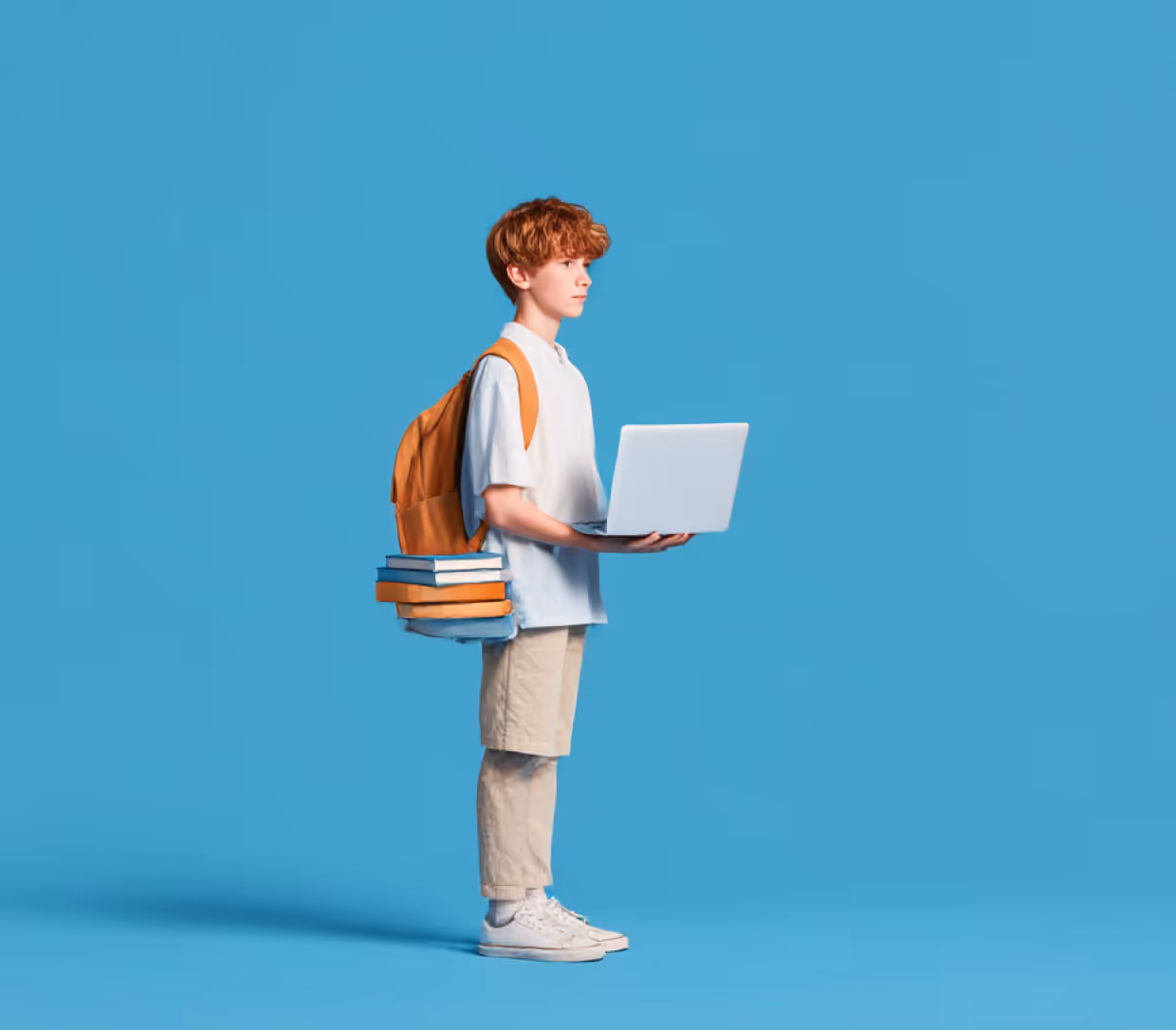 Підліток із рудим волоссям стоїть на синьому фоні з рюкзаком, книгами та відкритим ноутбуком у руках.