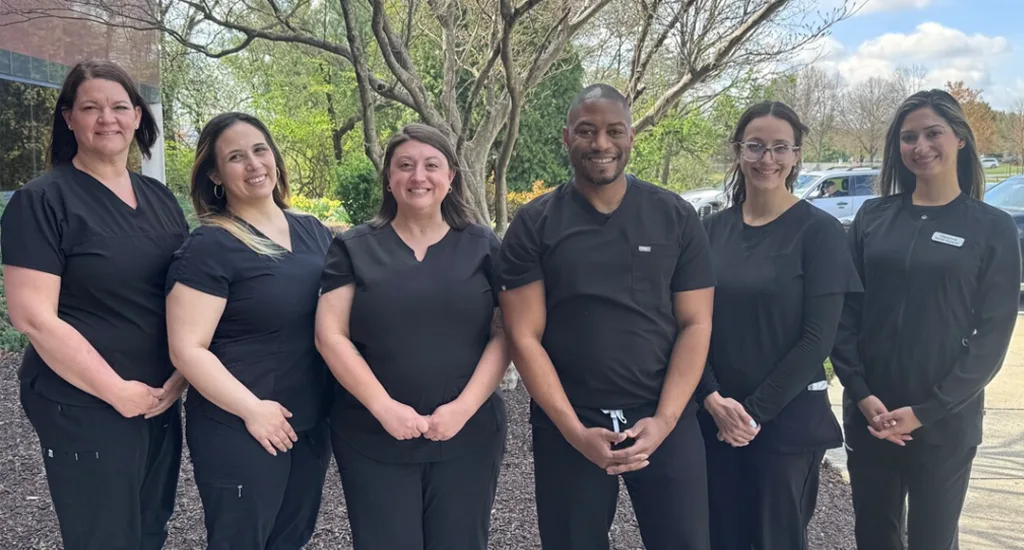 Group of smiling dental team members wearing black scrubs posing inside a clinic with Invisalign poster in the background.