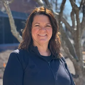 Young woman with long brown hair, glasses, and black top smiling against a plain background.
