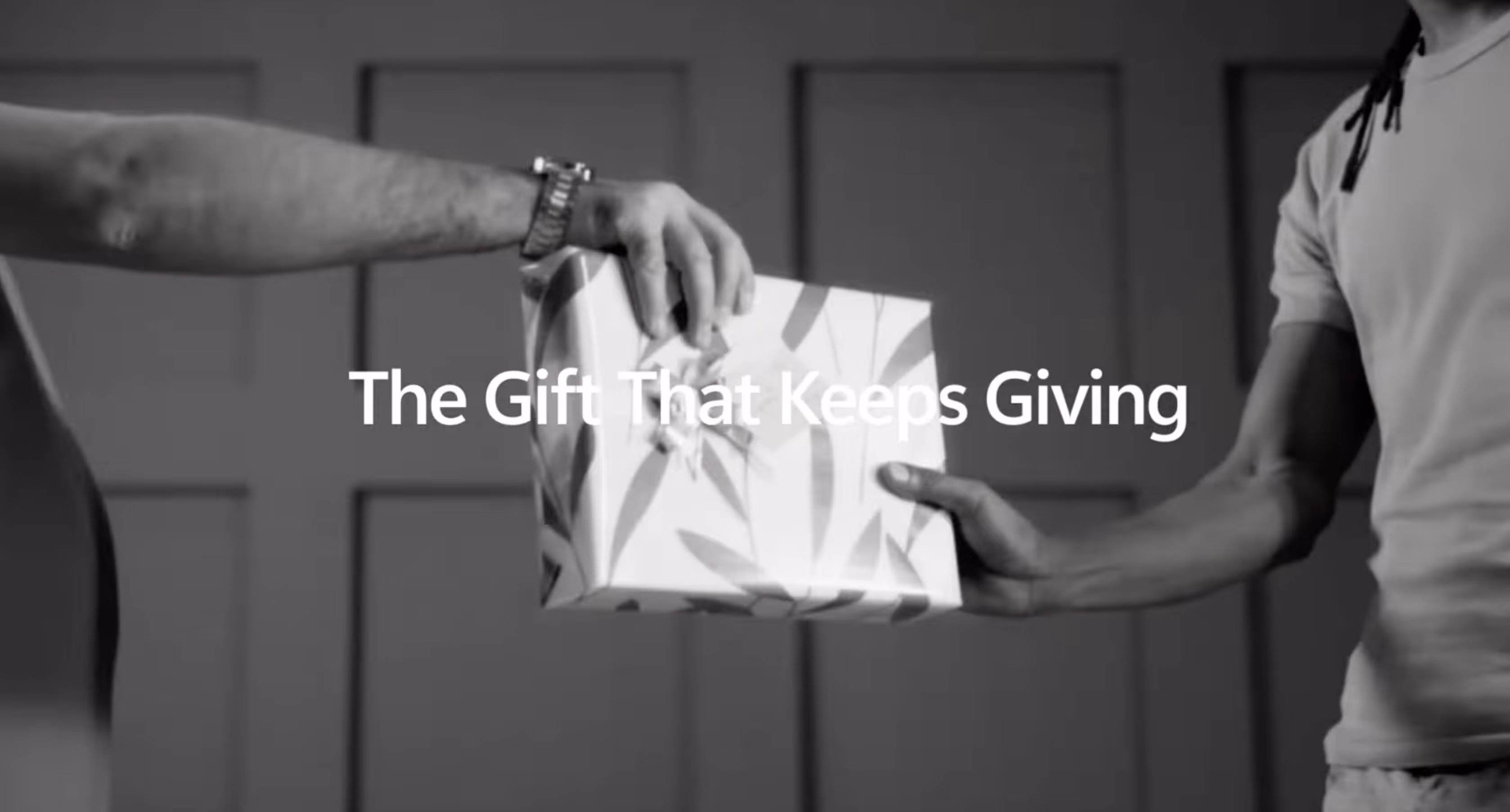 Two people exchanging a wrapped gift box with text overlay reading 'The Gift That Keeps Giving'.