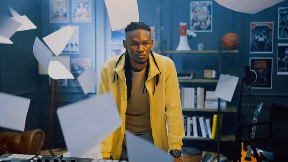 Man in a yellow jacket standing at a desk with papers flying around in a room with bookshelves, posters, and a basketball.