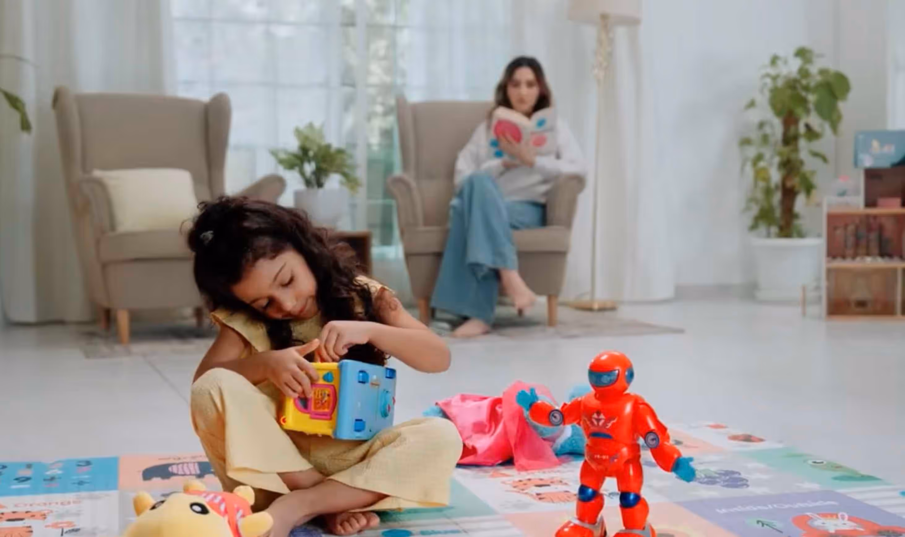 A young girl sitting cross-legged on a colorful play mat, playing with a multi-sided activity cube, with a red toy robot nearby and a woman reading a book in an armchair in the background.
