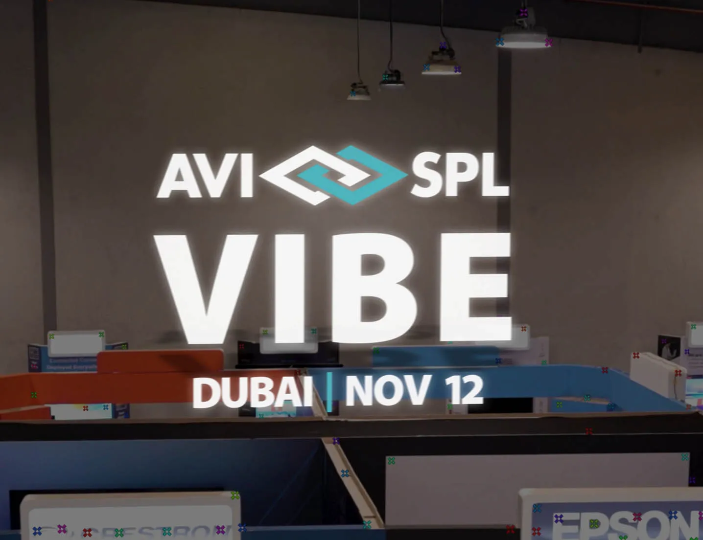 Projection on wall showing text AVI SPL VIBE Dubai Nov 12 in an office space with desks and pendant lights.