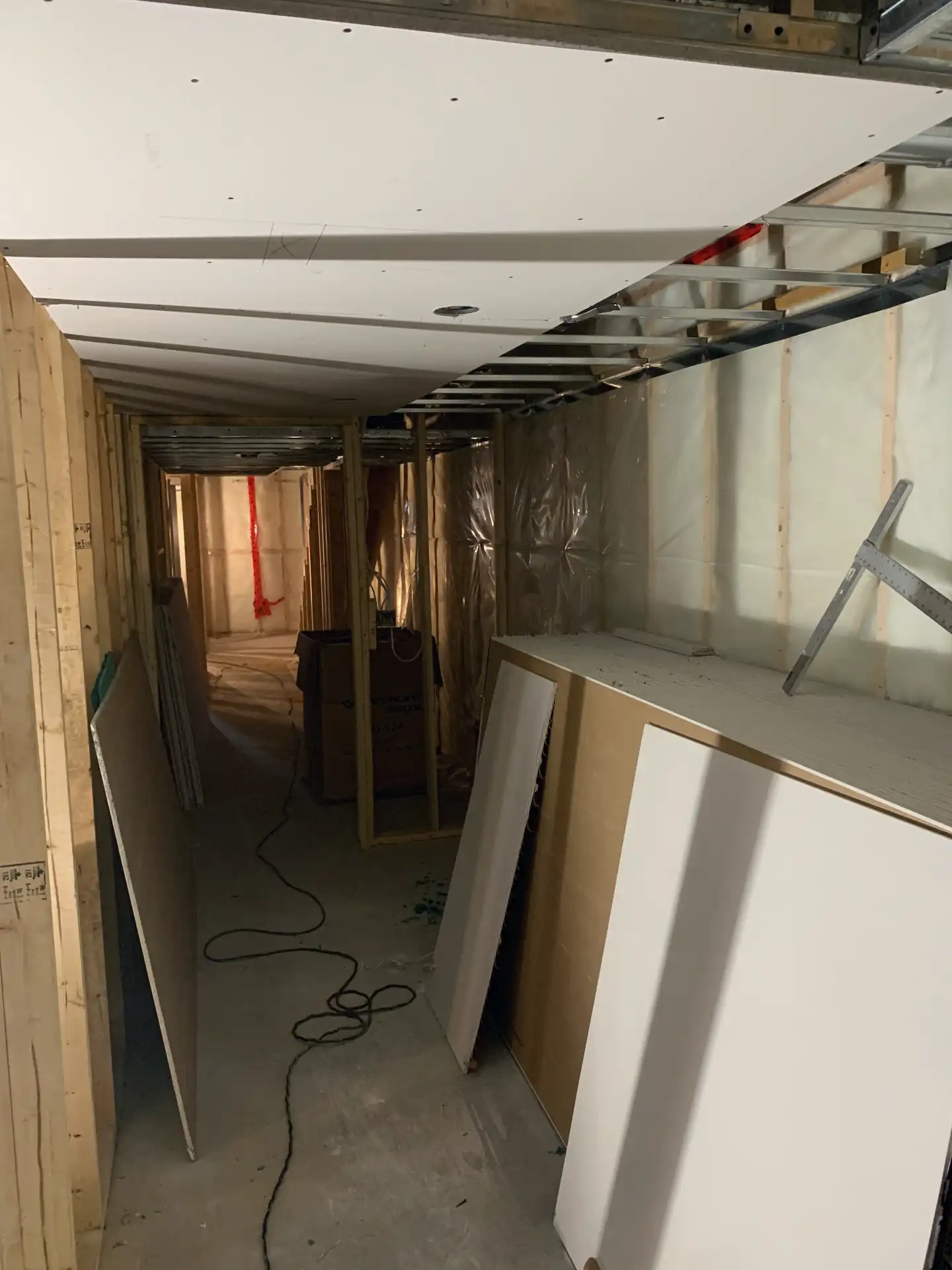 Drywall installation in basement hallway with ceiling boards being installed, framed walls, and vapor barrier in residential renovation.