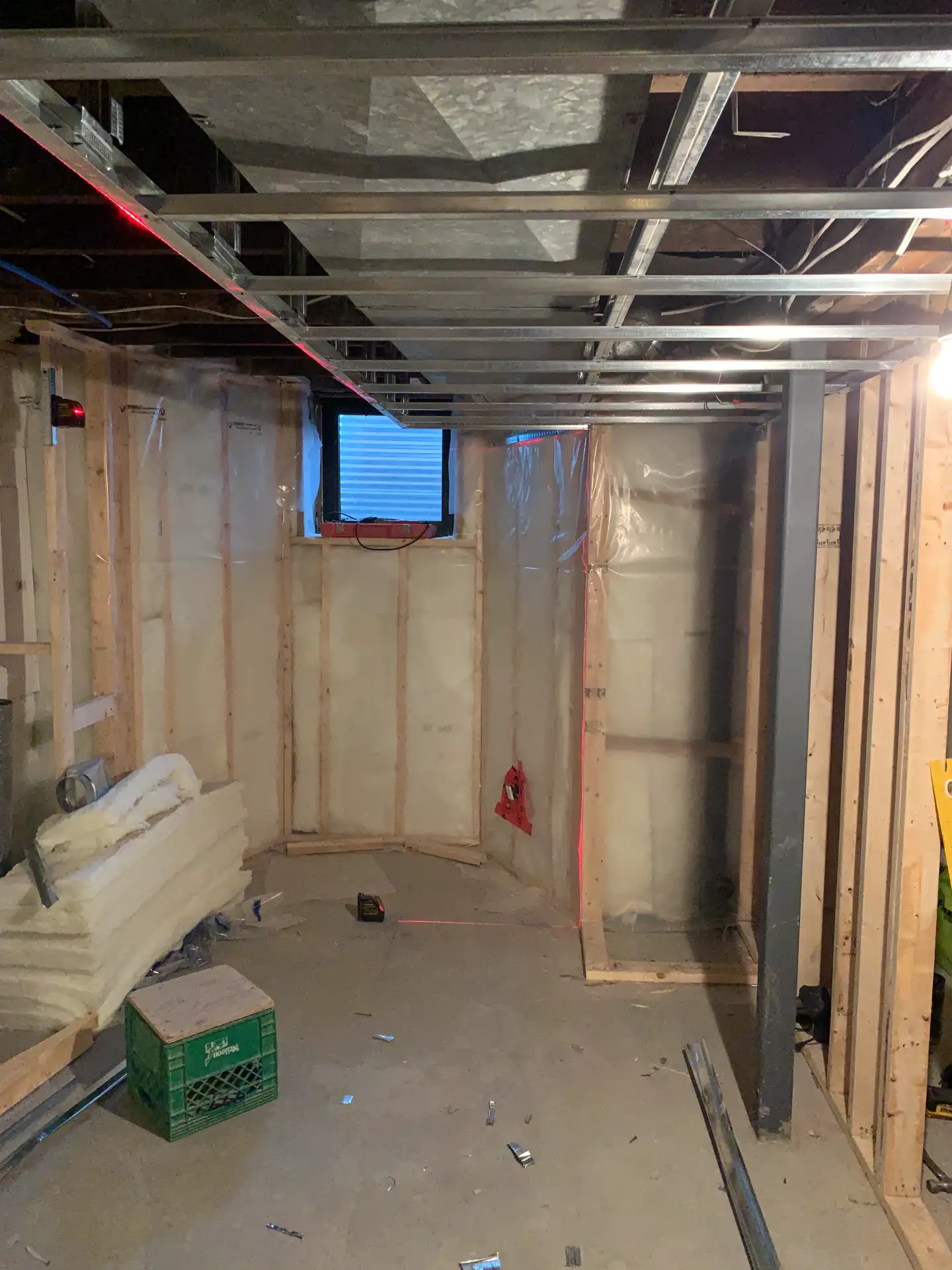 Unfinished basement with insulation, metal ceiling framework, exposed wooden studs, a small window, and construction materials on the floor.
